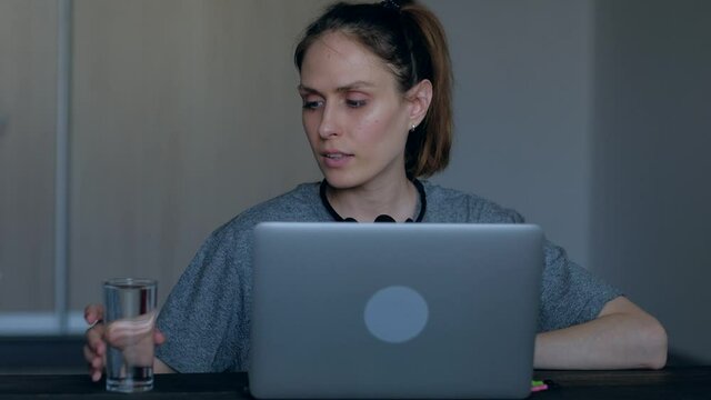 Female customer service representative working in home talking with client during the quarantine coronavirus period use headset, she is tired and takes a break to drink water from a glass . 4k.