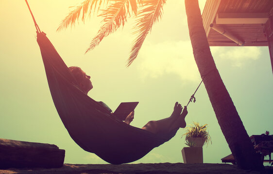 Woman an using a digital tablet  while relaxing in a hammock on a sand tropical summer beach