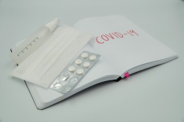 Covid 19 writing on a blank notebook with medicine pills, surgical face mask and hand sanitizer over white background