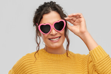 summer, valentine's day and eyewear concept - portrait of happy smiling young woman with pierced nose in pink heart-shaped sunglasses over grey background
