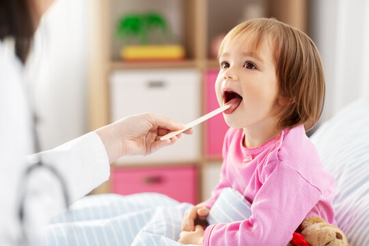 healthcare, medicine and people concept - doctor checking sick little girl's throat with tongue depressor in bed at home