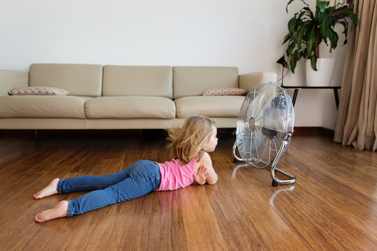 Little girl lying by electric fan on hardwood floor at home