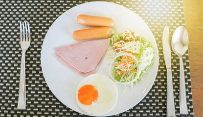 Breakfast has ham, fried egg, salad on a white plate.