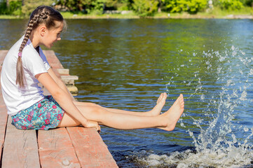 A girl in a white T-shirt sits by the lake on wooden walkways and splashes her bare feet in the water.