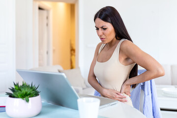 Businesswoman Holding Her Back While Working On Laptop At Home Office Desk. young woman, stretching, sitting feeling her back tired after working at laptop, small home office interior.