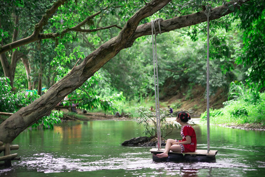 Beautiful happy girl, young pretty cheerful woman traveler sitting, swinging on a swing tied to a tree above the water, smiling in sunny day. Summer vacation, travel concept