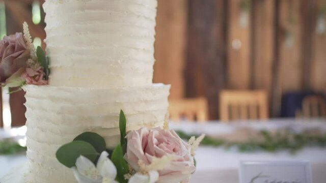 White wedding cake with flowers and texture frosting in barn on white table with love cake topper and shafts of sunlight- slow orbiting close up high key
