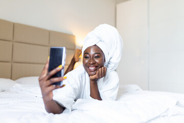 Smiling african young woman lying on bed in bathrobe with her mobile phone taking a selfie. African american woman relaxing on the bed after bath and looking at the phone camera taking a selfie