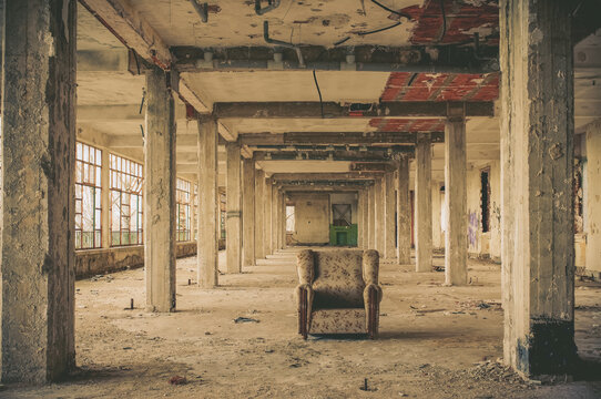 Lonely armchair inside abandoned industrial warehouse with decayed architecture and dramatic perspective. Urban exploration and post apocalyptic atmosphere concept.