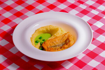 Fried fish with mashed potatoes and sauce served on a white plate over red plaid tablecloth.