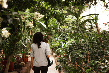 A black-haired young woman walks in the garden. A girl in a shirt walks through the greenhouse.