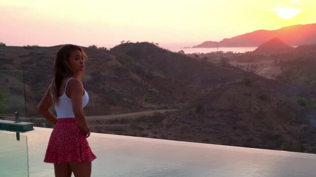 Slow motion shot of young woman standing by infinity pool while looking at sunset over sea - Bali, Kuta