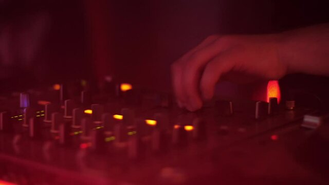DJ hands in a nightclub colored lights. Color lights show in nightclub. Hands of DJ tweak various track controls on DJ mixer console at nightclub. Fingers touch buttons sliders. Sound beat maker. 