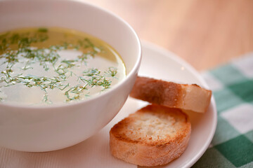 Chicken soup with vegetables and meat served in a white bowl over rustic wooden background with green plaid tablecloth.