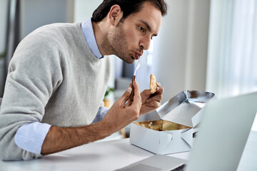 Young businessman eating while reading an e-mail on laptop at home.