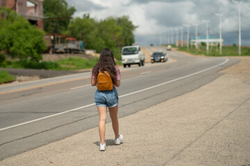 Girl tourists with backpacks hitchhiking on road. Hitchhiking tourism concept. Travel hitchhiker girl walking on road.  Girl in the pink t-shirt and denim shorts walking near the road.
