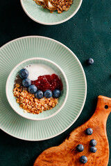 Top view of breakfast with granola, yoghurt, strawberry jam, chia seeds and blueberry on a dark green table.