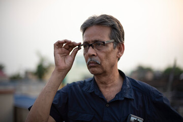 An old / aged Indian Bengali man in blue shirt is adjusting his spectacles while standing on a rooftop under the open sky. Indian lifestyle and seniors