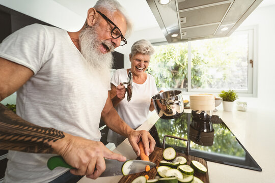 Happy senior couple having fun cooking together at home - Elderly people preparing health lunch in modern kitchen - Retired lifestyle family time and food nutrition concept