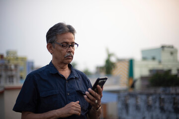 An old / aged Indian Bengali man in blue shirt is watching his cellphone while standing on a rooftop under the open sky. Indian lifestyle and seniors