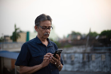 An old / aged Indian Bengali man in blue shirt is watching his cellphone while standing on a rooftop under the open sky. Indian lifestyle and seniors