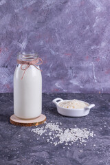 Vegan rice milk in a glass bottle and rice in a white plate on a wooden stand on a gray background with a gray napkin