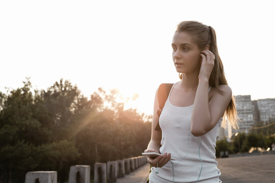 Beauty young female model with long blond hairstyle in sport clothes and with backpack standing on the road and looking sideways. The girl listen to music in earphones and walking on the cort