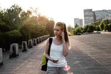 Beauty young female model with long blond hairstyle in sport clothes and with backpack standing on the road and looking sideways. The girl listen to music in earphones and walking on the cort