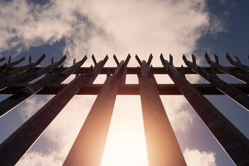 Metal industrial security fence with spikes on top against blue sky background. Concept safety, protection of property perimiter. Sun flare. - obrazy, fototapety, plakaty