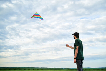 Jun 5, 2020-Ternopil/Ukraine:Young brunette skinny man, wearing dark green t-shirt, playing with colorful kite on green field meadow in summer. Kite flying in blue cloudy sky. Family leisure activity