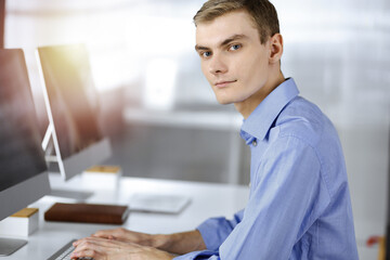 Fototapeta premium Young successful businessman works with computer, sitting at the desk in a sunny modern office. Headshot of male entrepreneur or it-specialist at workplace. Business concept