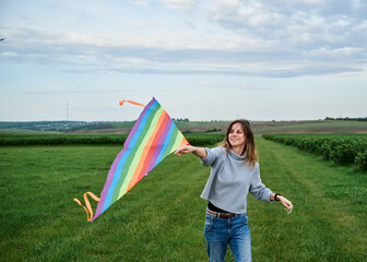 Young brunette woman, wearing casual clothes green t-shirt, playing with colorful kite on green field meadow in summer, running, jumping. Family leisure activity at natural rural landscape.