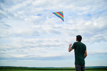 Jun 5, 2020-Ternopil/Ukraine:Young brunette skinny man, wearing dark green t-shirt, playing with colorful kite on green field meadow in summer. Kite flying in blue cloudy sky. Family leisure activity