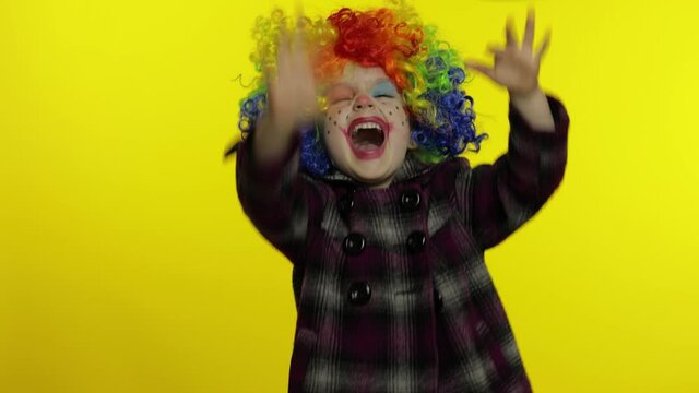Little child girl clown in colorful wig hides behind her hands and shows funny faces. Halloween