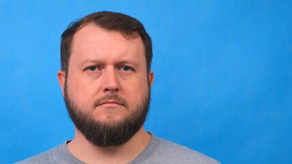 Portrait of a sad bearded man in a gray t-shirt singing and talking to the camera on a blue background.