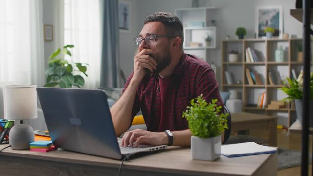 Tired Man In Red Shirt Yawning And Using Computer For Remote Work At Home.