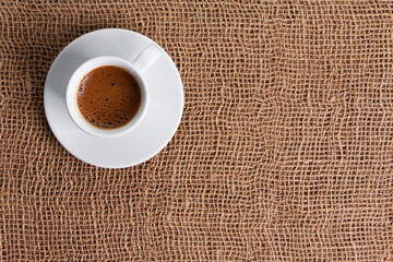 Single white cup of black coffee with foam on jute burlap sackcloth. Top view, sack copy space. Coffee shop, morning, agriculture, traditional roast concept
