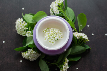 Lilac jar with white cream of delicate texture, inside which-white flowers. Spirea twigs with flowers and green leaves are lying around. Dark background 