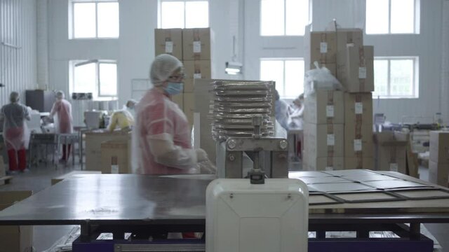 Caucasian woman putting foil sheet in die cutting machine and collecting cutted covers for food dish containers. Wide shot of factory manufacturing packaging. Conveyor, business, production industry.