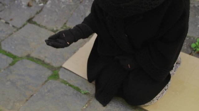 A beggar in a black robe with a hood begs for alms in the street