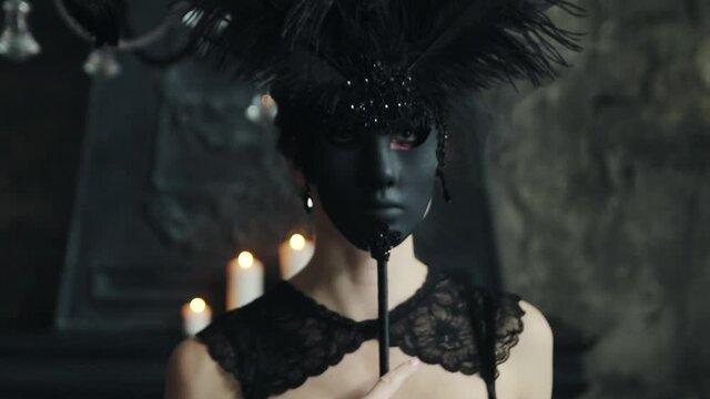 A girl in black lace underwear and a black masquerade mask with feathers walks around the room