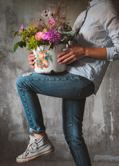 Stylish Woman jeans shirt professional florist holds composition wild flowers pot Flower shop Background, concrete, gray wall.
