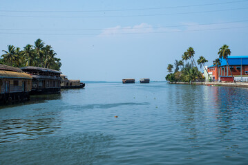 Fototapeta na wymiar Small houses in a local village located next to Kerala's backwater on a bright sunny day and traditional Houseboat seen sailing through the picturesque backwaters of Allapuzza or Alleppey in Kerala 