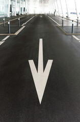 White arrow street sign on dark asphalt, pointing downwards at a parking zone with futuristic white architecture columns in the blurry background.