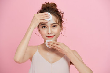 Close up beauty portrait of a smiling attractive woman cleaning her face with a cotton pad isolated over pink background