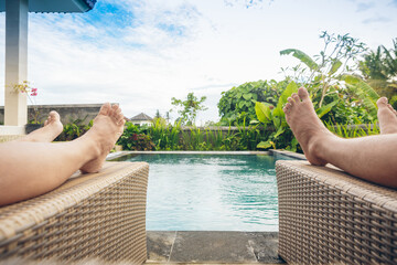 Sunbathing on the pool bed near clear blue water in the swimming pool at tropical resort near beach in summer day. Feet of couple