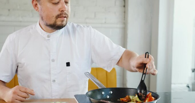 Young chef cooking vegetables, frying in pan colorful pieces, mixing with spoon