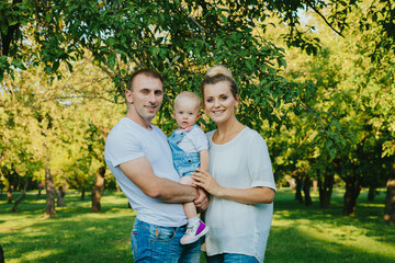 Fototapeta premium Happy young family on nature background. Smiling father and mother holding their little child's in his arms. Young man, woman and child in park on summer. Love and harmony in family.