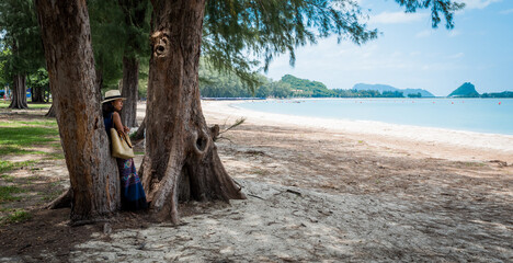 Woman with white fedora and sarong leaning against tree at the beach looking at the sea