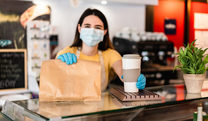 Young woman wearing face mask while serving takeaway breakfast and coffee inside cafeteria restaurant - Worker preparing healthy food inside cafè bar during coronavirus period - Focus on right hand